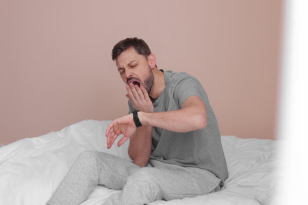 Sleepy man checking time on watch in bed at homeの写真素材