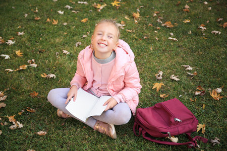 Cute little girl with copybook and backpack on green grass outdoors