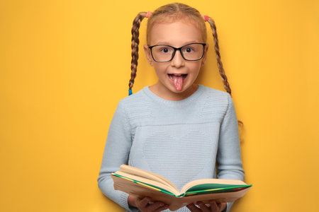 Cute little girl with glasses and textbook on yellow background