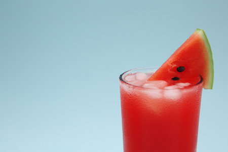 Delicious drink with ice cubes and piece of fresh watermelon on light blue background, closeup. Space for textの写真素材