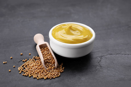 Bowl of delicious mustard and scoop with seeds on black textured table