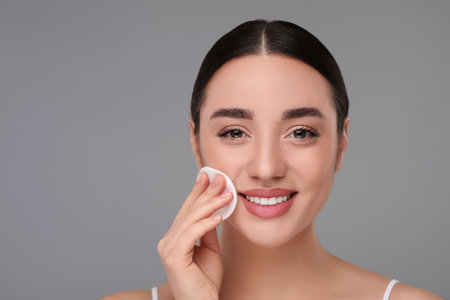 Beautiful woman removing makeup with cotton pad on gray background, space for textの写真素材