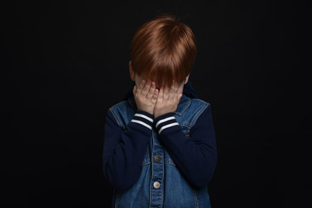 Boy covering face with hands on black background. Children's bullyingの写真素材