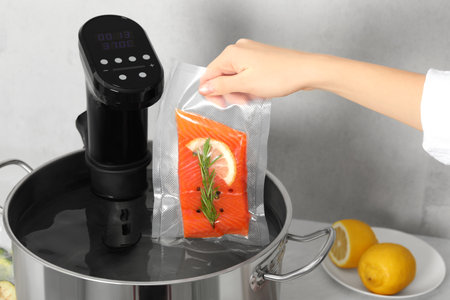 Woman putting vacuum packed salmon into pot with sous vide cooker in kitchen, closeup. Thermal immersion circulatorの写真素材
