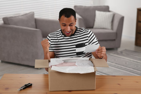 Emotional young man with greeting card near parcel at table indoors. Internet shopping