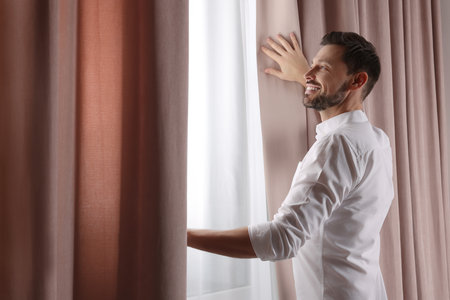 Happy man opening window curtains at home. Space for textの写真素材