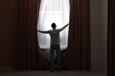Man opening window curtains at home, back viewの写真素材