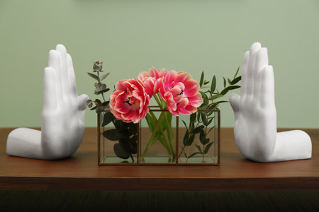 Glass holder with green branches and beautiful flowers near hand shaped bookends on wooden table indoors. Stylish interior designの写真素材