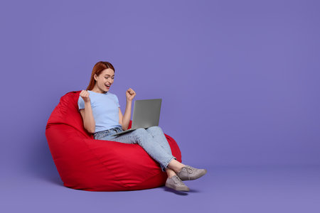 Happy young woman with laptop sitting on beanbag chair against lilac background, space for textの写真素材