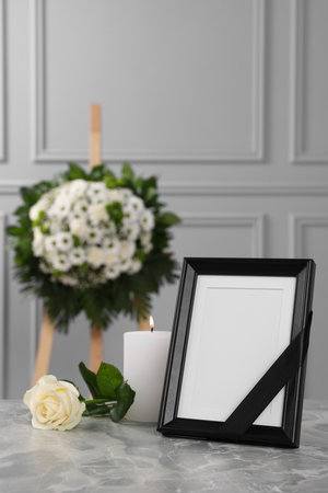 Photo frame with black ribbon, rose, burning candle on light gray table and wreath of flowers near wall indoors. Funeral attributesの写真素材