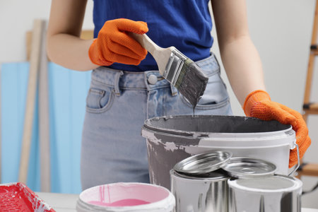 Woman dipping brush into bucket of gray paint at table indoors, closeup