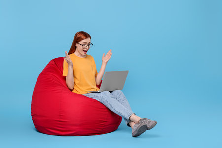 Surprised young woman with laptop sitting on beanbag chair against light blue background, space for textの写真素材