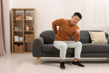 Asian man suffering from back pain on sofa indoorsの写真素材