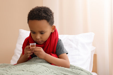 African-American boy taking cough syrup on bed at home, space for text. Cold medicineの写真素材