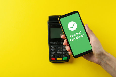 Woman using terminal for contactless payment with smartphone on yellow background, top view....