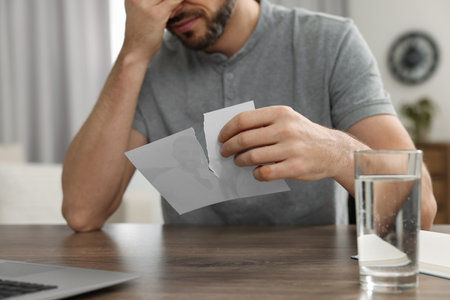 Man holding torn photo at table indoors, closeup. Divorce concept