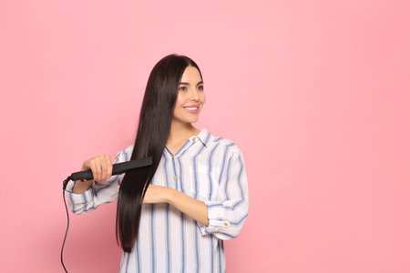 Beautiful happy woman using hair iron on pink background. Space for textの写真素材