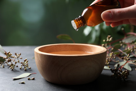 Woman dripping eucalyptus essential oil from bottle into bowl at gray table, closeupの写真素材