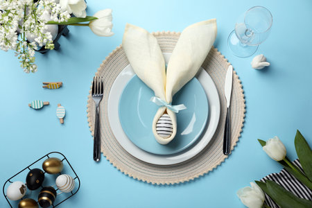 Festive table setting with painted eggs, plates and white tulips on light blue background, flat lay....