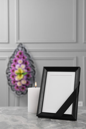 Photo frame with black ribbon, burning candle on light gray table and wreath of plastic flowers near wall indoors. Funeral attributesの写真素材