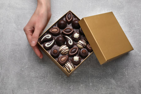 Woman with open box of delicious chocolate candies at light gray table, top view