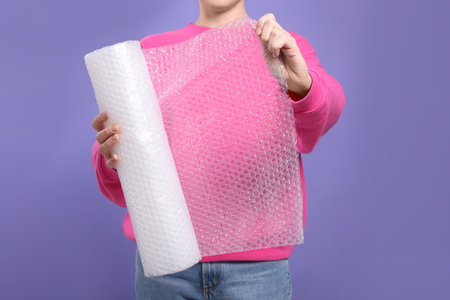Woman holding roll of bubble plastic wrap on purple background, closeup. Stress relief