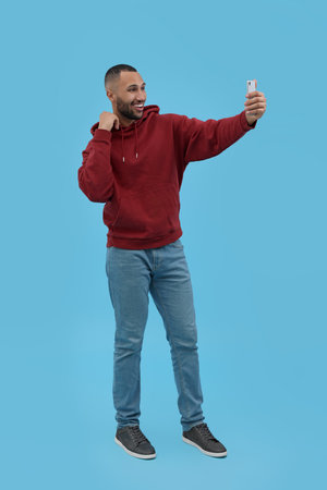 Smiling young man taking selfie with smartphone on light blue background