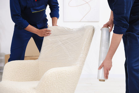 Male movers with stretch film wrapping armchair in house, closeupの写真素材
