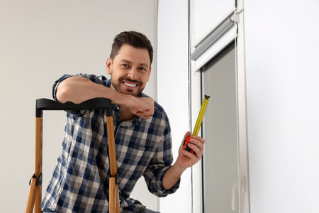 Man with measure tape on stepladder near window indoors. Roller blinds installationの写真素材