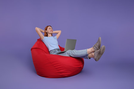 Happy young woman with laptop sitting on beanbag chair against lilac backgroundの写真素材