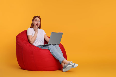 Surprised young woman with laptop sitting on beanbag chair against yellow backgroundの写真素材