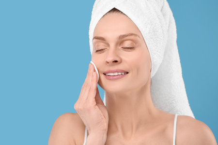 Beautiful woman in terry towel removing makeup with cotton pad on light blue background, space for textの写真素材