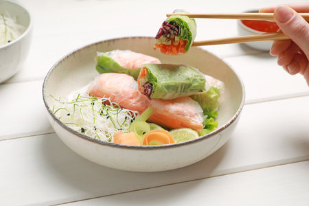 Woman eating delicious spring rolls wrapped in rice paper with chopsticks at white wooden table, closeupの写真素材