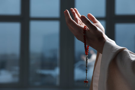 Muslim man with misbaha praying near window indoors, closeup. Space for text