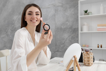Beautiful young woman applying blusher with brush at dressing table indoorsの写真素材