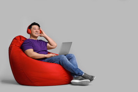 Smiling man with laptop and headphones sitting in beanbag chair against grey backgroundの写真素材
