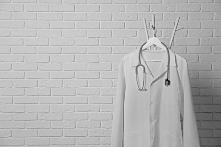 Medical uniform and stethoscope hanging on rack near white brick wall. Space for text