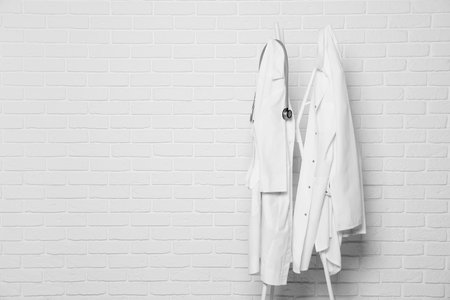 Medical uniforms and stethoscope hanging on rack near white brick wall. Space for textの写真素材