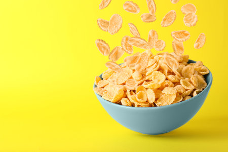 Tasty corn flakes falling into bowl on yellow background, space for text