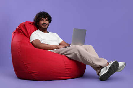 Smiling man with laptop sitting on beanbag chair against purple backgroundの写真素材