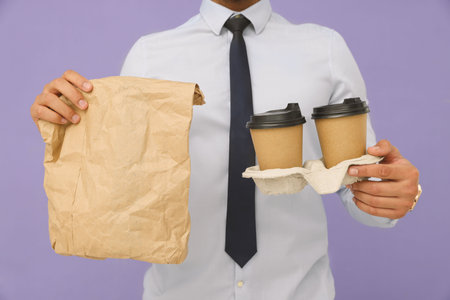 Young intern holding takeaway cups with hot drink and paper bag on lilac background, closeupの写真素材