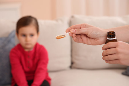 Mother with cough syrup for her daughter in room, focus on handsの写真素材