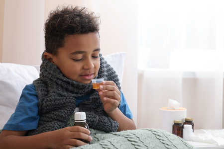 African-American boy taking cough syrup on bed at home, space for text. Cold medicineの写真素材