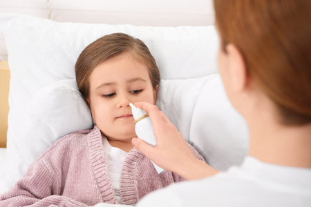 Mother using nasal spray to treat her little daughter on bedの写真素材