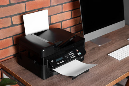 Modern printer with paper near computer on wooden desk at home