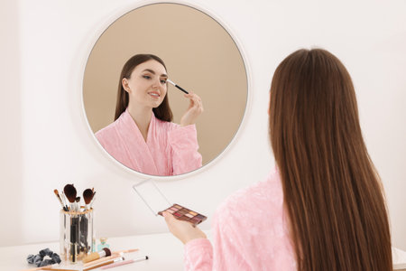 Beautiful young woman applying eyeshadow with brush near mirror indoorsの写真素材