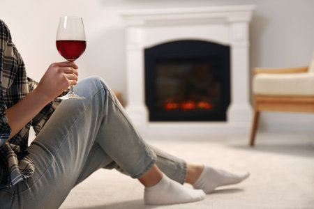 Young woman with glass of wine resting near fireplace at home, closeupの写真素材