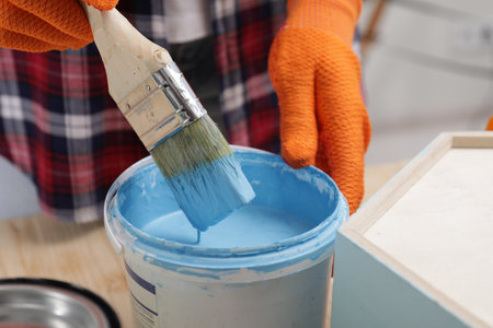 Woman dipping brush into bucket of light blue paint at wooden table indoors, closeupの写真素材