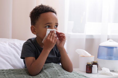 African-American boy with tissue blowing nose in bed indoors, space for text. Cold symptomsの写真素材