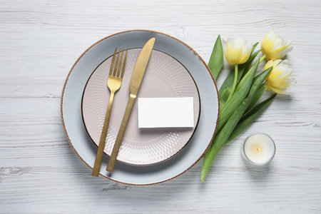 Stylish table setting with cutlery, tulips and scented candle on white wooden background, flat lay